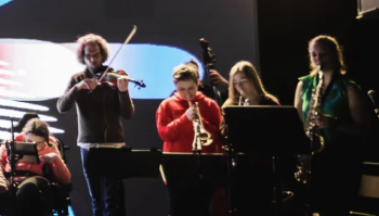 A diverse group of musicians, including a violinist, clarinetist, saxophonist, and a person in a wheelchair using assistive technology, performing live on stage against an abstract digital backdrop.