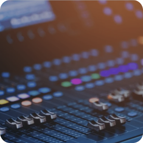 A close-up of a professional mixing console with illuminated faders, colourful buttons, and control knobs, with a warm ambient lighting effect.
